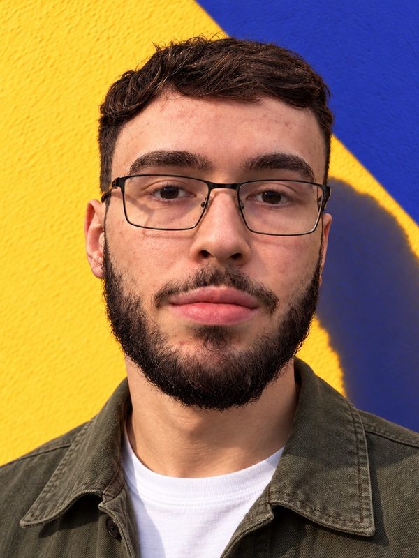 Natã, de óculos e barba, com jaqueta verde-oliva, diante de uma parede amarela.