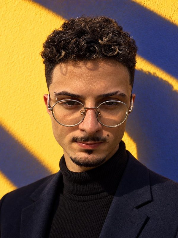 Victor, de óculos redondos e cabelo cacheado, com blazer escuro, diante de uma parede amarela.